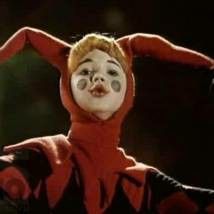 a clown with red hair and white face paint on it's head, holding his arms in the air