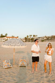 a man and woman standing in the sand under an umbrella at the beach with chairs