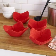 three red bowls on a wooden counter with a black spoon in the middle and a pot holder next to them