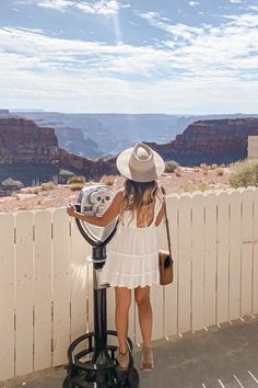 a woman in a white dress and hat standing next to a parking meter at the edge of a cliff