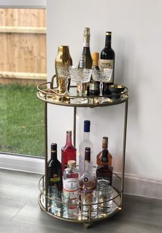 a gold bar cart with liquor bottles and glasses on the top shelf in front of a window