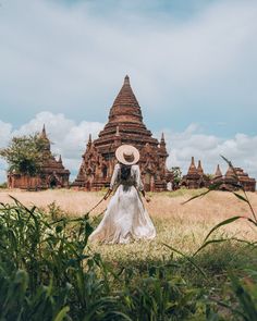 a woman in a white dress and straw hat walks through the grass towards an old building