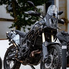 a black motorcycle parked on top of a sidewalk next to a tree and building in the background