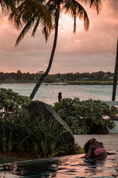 two people are sitting in the water next to some palm trees and bushes at sunset