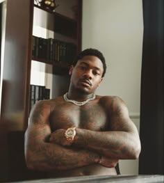 a shirtless man with his arms crossed standing in front of a bookshelf