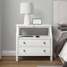 a night stand with two drawers and a lamp on it next to a white bed