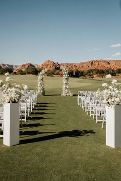 an outdoor ceremony setup with white chairs and flowers on the grass in front of mountains