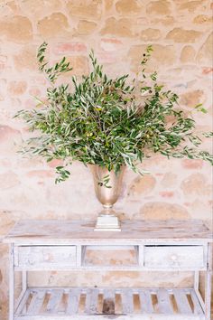 a vase filled with green plants sitting on top of a white wooden table next to a brick wall