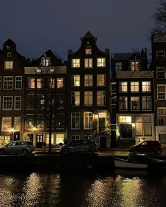 some cars parked on the side of a body of water at night with buildings in the background