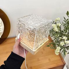 a person is holding a glass block on a table next to flowers and a clock