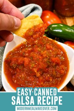 a hand holding a tortilla chip over a bowl of salsa sauce with peppers and jalapenos in the background