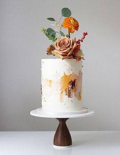 a white cake with orange and yellow flowers sitting on top of a wooden stand in front of a gray wall