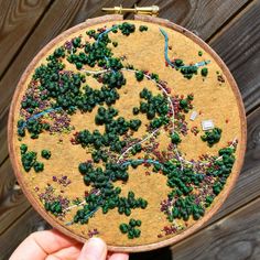 a person holding up a small embroidery piece with green trees on it and other things in the background