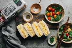 an assortment of food on a cutting board with chopsticks next to it and two bowls filled with vegetables