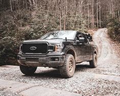 a black truck driving down a dirt road in front of some trees and bushes with no leaves on it