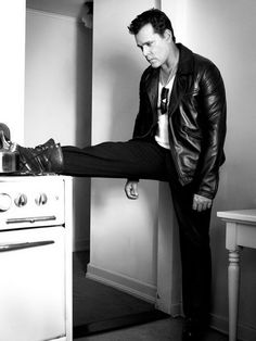 black and white photograph of man leaning on stove with leg in air, wearing leather jacket