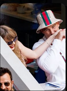 a man in a hat and sunglasses leaning against a wall next to a woman with her hand on her face