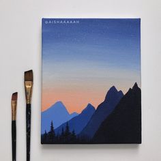 two brushes are next to a painting on a white surface with mountains in the background