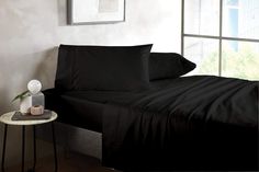 a bed with black sheets and pillows next to a small table in front of a window