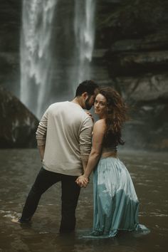 a man and woman are standing in the water near a waterfall holding each other's hands