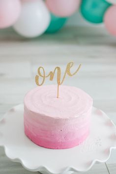 a pink and gold cake with the word one on top sitting on a white plate