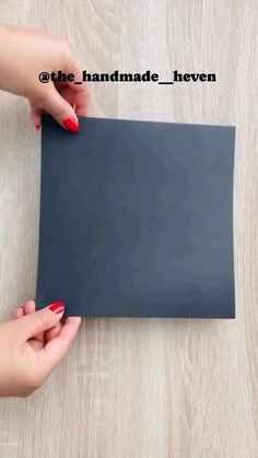 two hands holding a piece of black paper on top of a wooden table with red nail polish