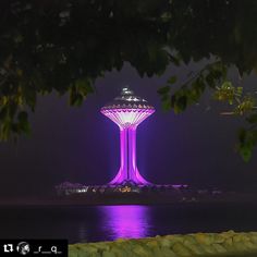 a purple light tower is lit up in the night sky over water and trees with lights on it