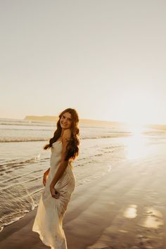 a woman standing on top of a sandy beach next to the ocean in a white dress