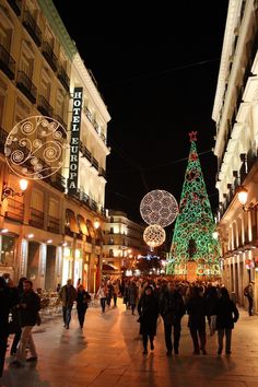 people are walking down the street in front of christmas trees and lights on buildings at night