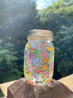 a painted mason jar sitting on top of a piece of wood with trees in the background