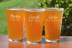 three glasses filled with beer sitting on top of a wooden table next to grass and trees