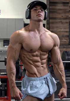 a shirtless man with headphones and no shirt standing in a gym holding a pair of dumbbells