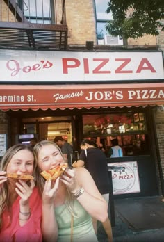 two young women eating pizza in front of a pizzeria on a city street