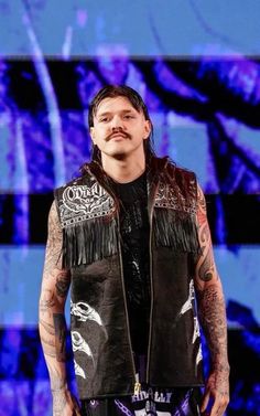 a man with long hair and tattoos standing in front of a blue background wearing a leather vest