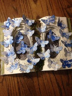 an open book with blue butterflies on it and moss in the middle, sitting on a wooden table