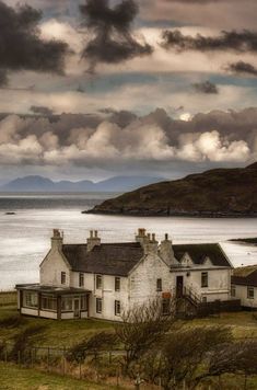 an old house sitting on top of a lush green field next to the ocean under a cloudy sky