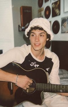 a young man sitting on top of a bed holding an acoustic guitar in his hand