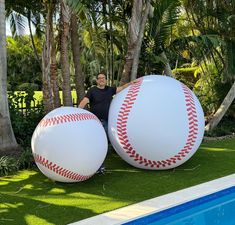 a man sitting next to two giant baseball balls in the grass near a swimming pool