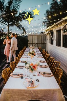 a long table set up for an outdoor dinner party with string lights strung over it