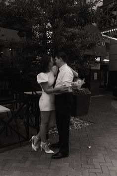 a man and woman kissing in front of a tree on a sidewalk with lights above them