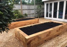 two wooden raised beds with black soil in front of a backyard patio and garden area