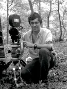 a man squatting down next to a camera in the woods with trees behind him