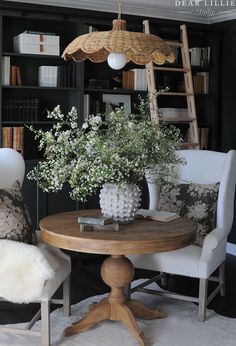 two chairs and a table with flowers in vases on it, near bookshelves