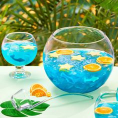 two glasses filled with blue liquid and orange slices on top of a table next to plants