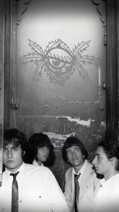 black and white photograph of four men wearing ties in front of a door with an eye drawn on it