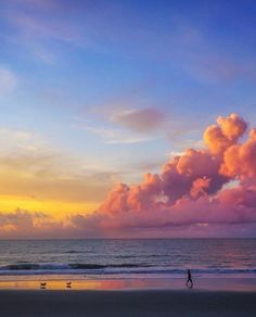 a person walking on the beach with a surfboard under a pink and blue sky