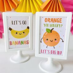 two framed pictures with oranges and banana's on them next to some paper fans