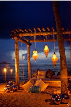 an outdoor seating area on the beach with lanterns hanging from it's posts and pillows