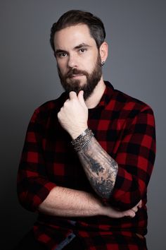 a man with a beard and tattoos on his arm is posing for a photo in front of a gray background