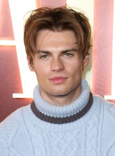 a young man wearing a blue sweater looks at the camera while standing in front of a red wall